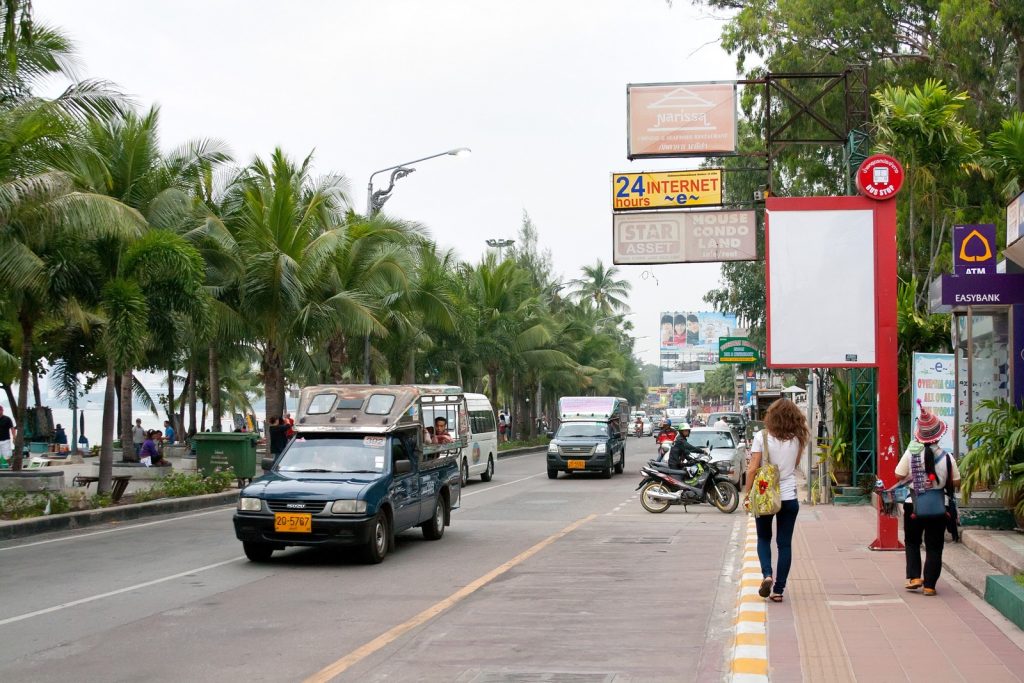 芭提雅海滩路上蓝色双条车与汽车摩托车混行，路旁可见棕榈树和人行道