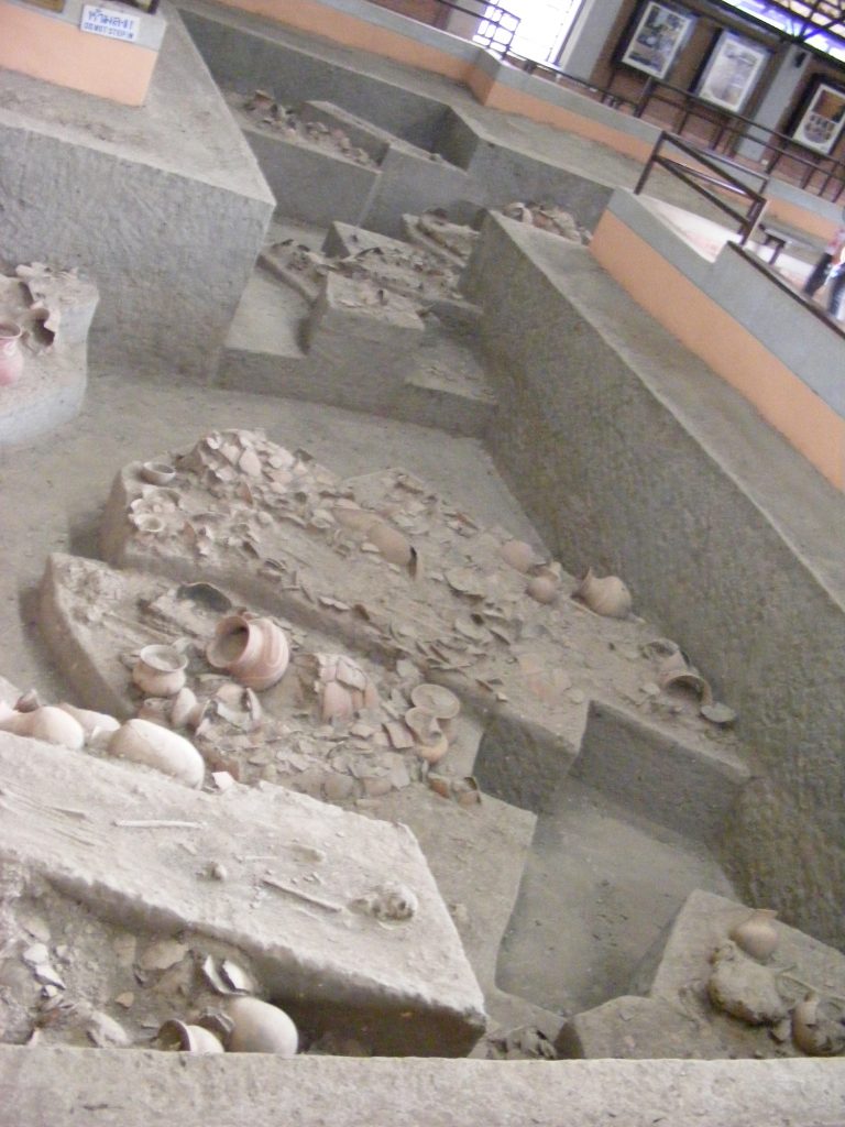 诗奈寺室内发掘展示俯视图，可见分层坑方、遗骸和陶器