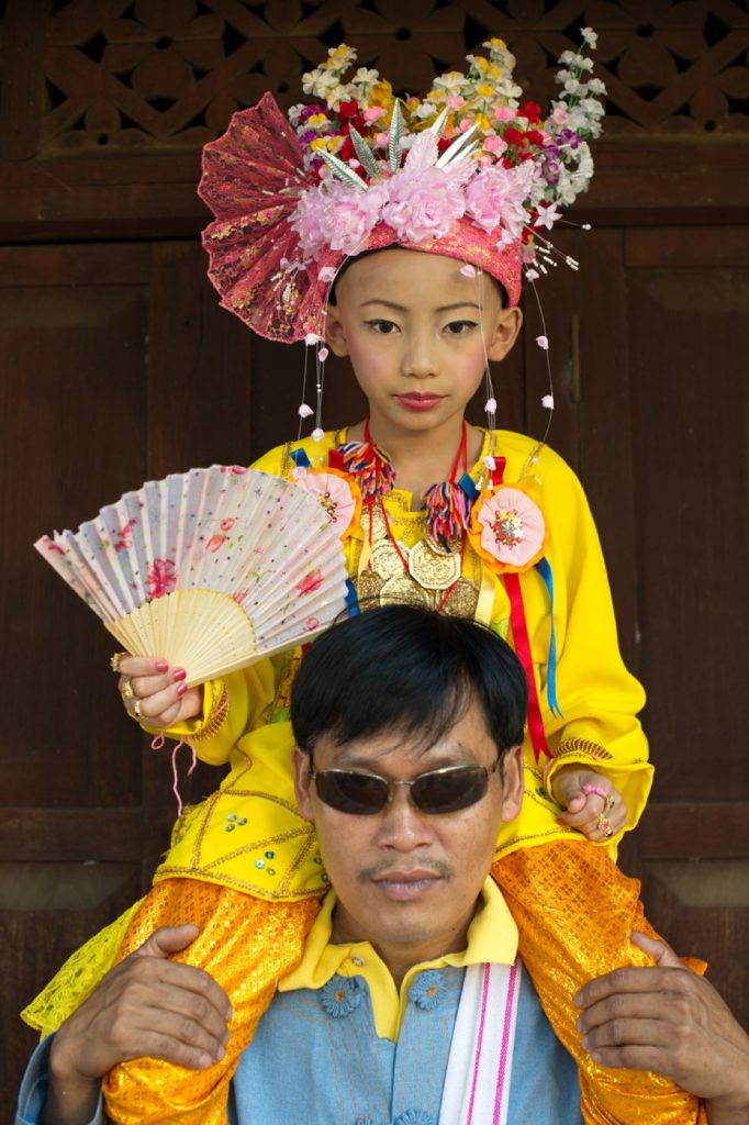佩戴花冠和扇子的盛装少年坐在一名成年男子肩上