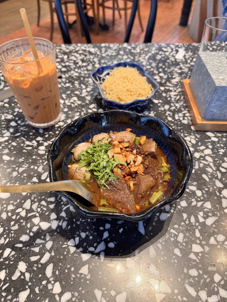 黑色花边碗里盛着深色船面，桌上另有饮料和一小碟面条