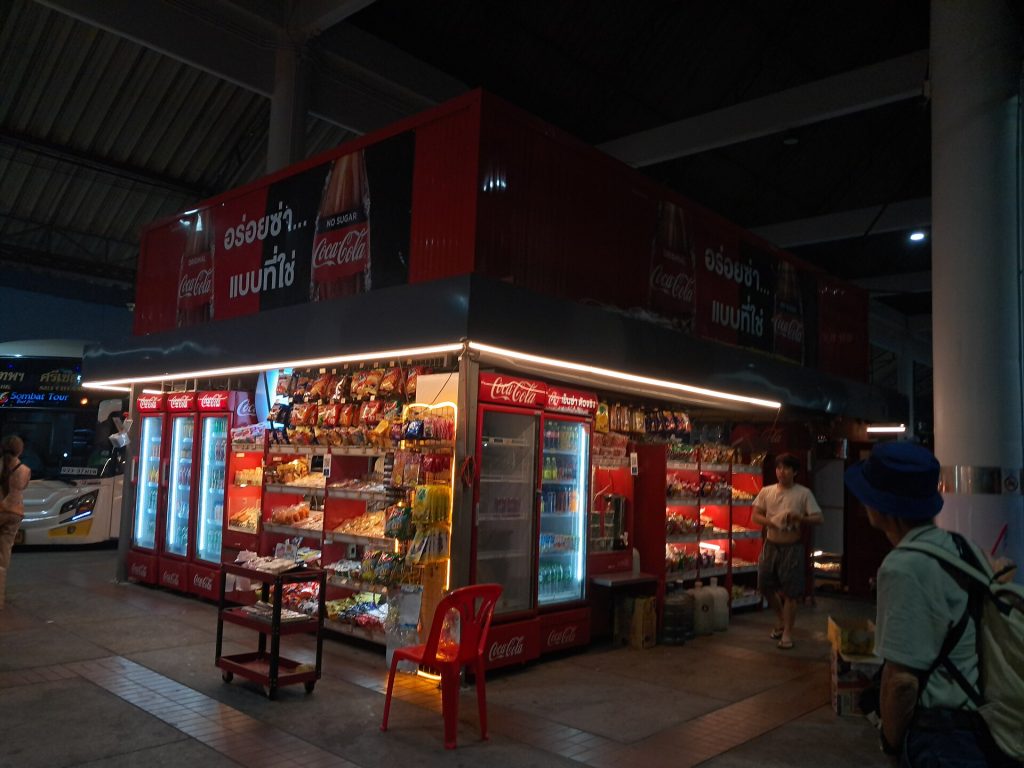 莫奇二号站台内亮灯的零食亭，旁边可见长途巴士车头与候车旅客