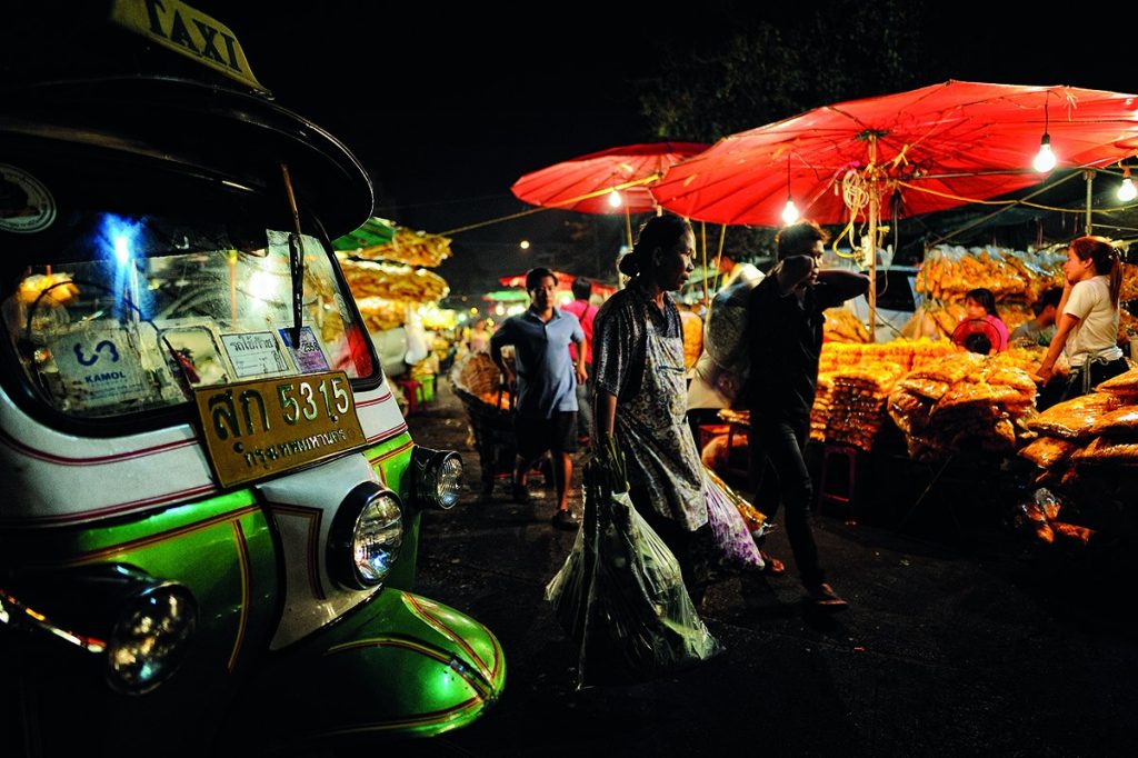 夜间街边红伞下是花摊，左侧停着嘟嘟车，行人从车与摊位之间经过。