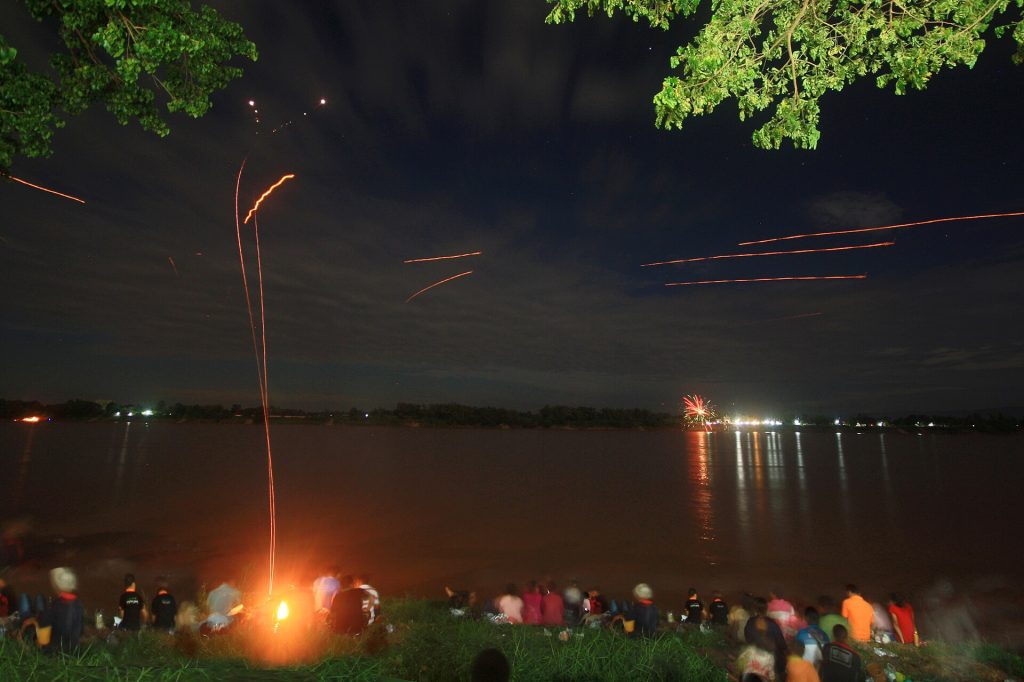 夜色中的湄公河岸聚集着观众，河面上方出现多条橙红色长曝光光轨