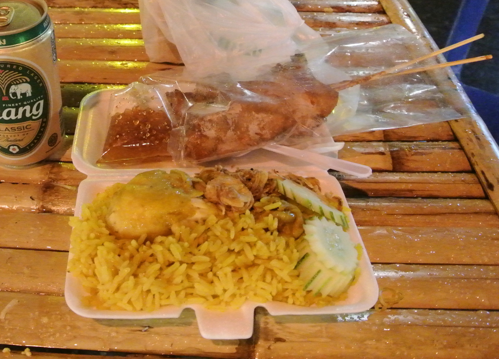泡沫餐盒里的鸡肉黄饭配鸡肉、黄瓜和炸红葱，旁边放着烤串