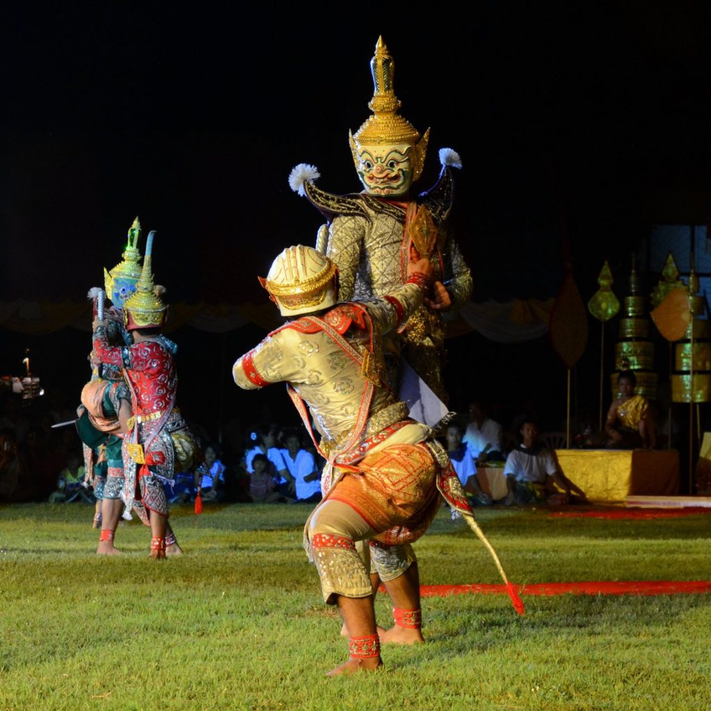 夜间草地上三名戴面具的孔剧演员表演对抗动作，前景演员屈膝转身，后方可见观众。