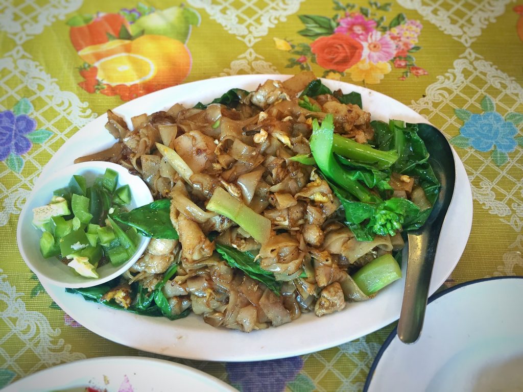 花色桌布上的炒粉全盘，棕色宽粉间有鸡肉、鸡蛋和芥蓝，一旁小碟装绿色辣椒。