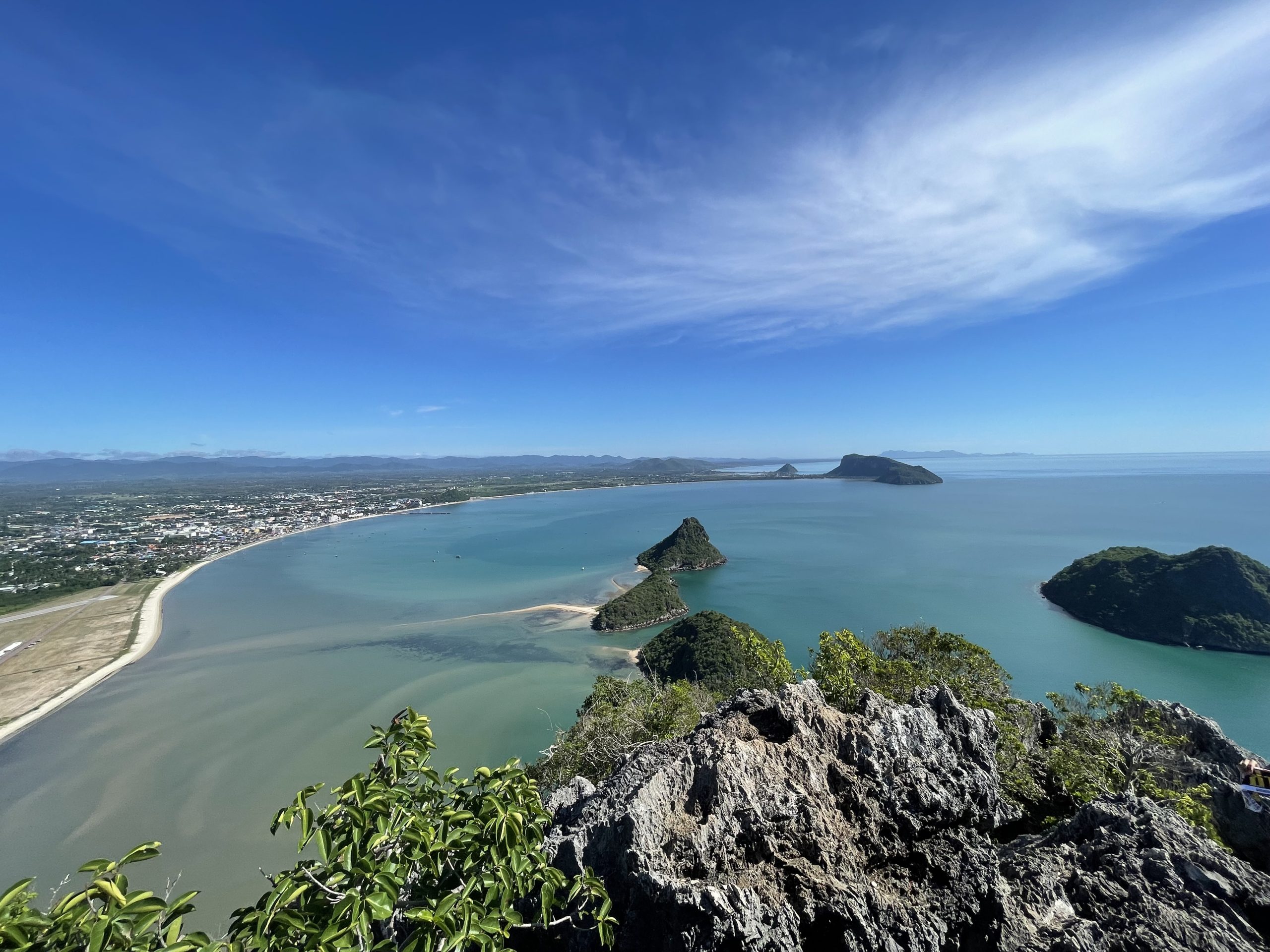 2024年Lom Muak山顶向北看相邻Ao Prachuap巴蜀湾,前景尖石绿叶,弧形城区海岸、海中丘体和浅色沙带;非Ao Manao本湾