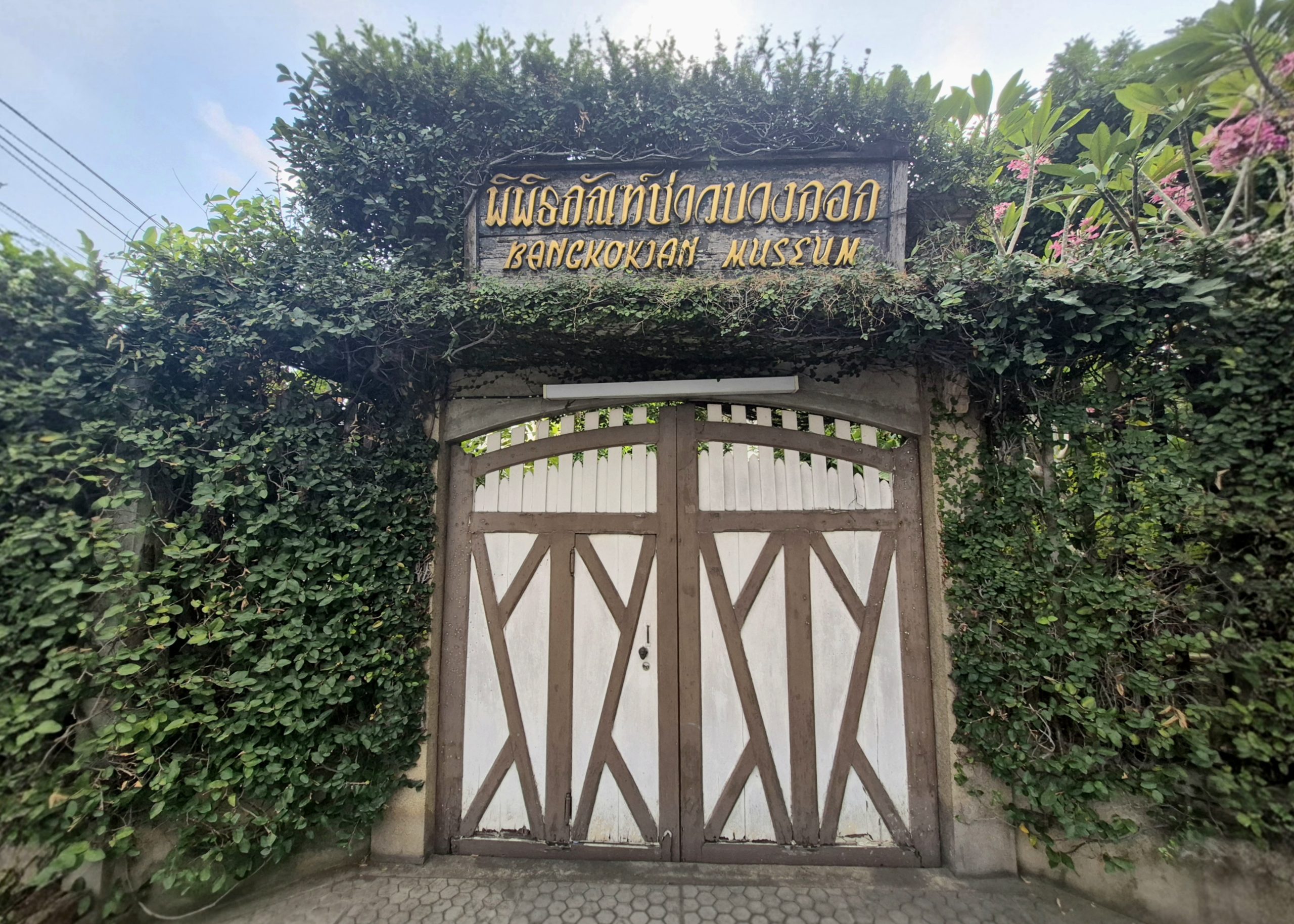 曼谷民俗博物馆双扇木门与泰英馆名门牌，墙上叶蔓和右上方粉红花，2026年2月拍摄