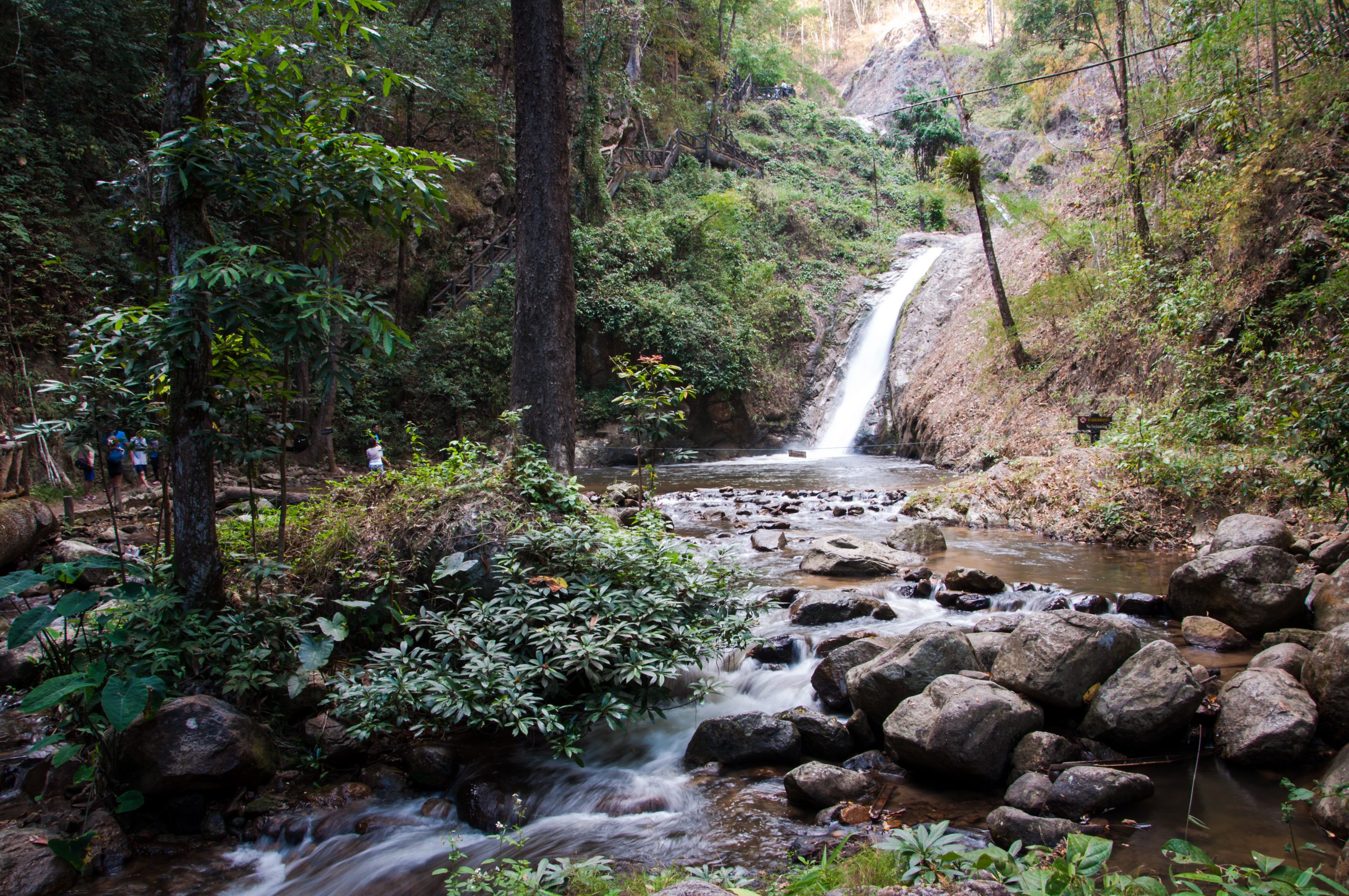 杰颂瀑布白色水流沿裸露岩面落入潭中,前景有流水石块和植物,左侧为林间步道栏杆