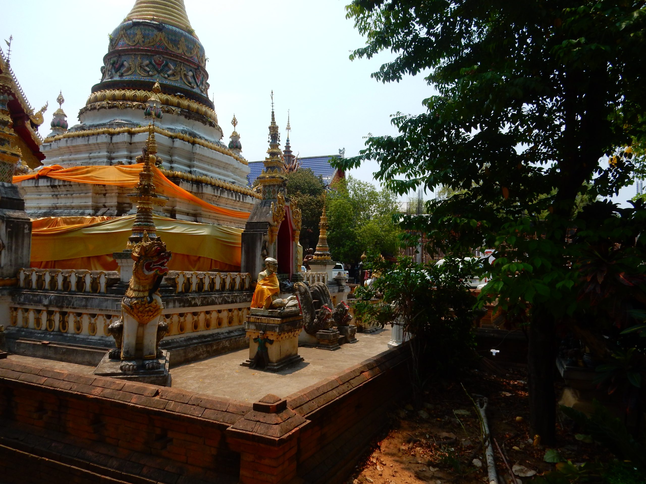 清迈 Wat Chai Mongkhon 佛塔的白色层叠基座与橙黄色围布，周围有尖顶构件和树木