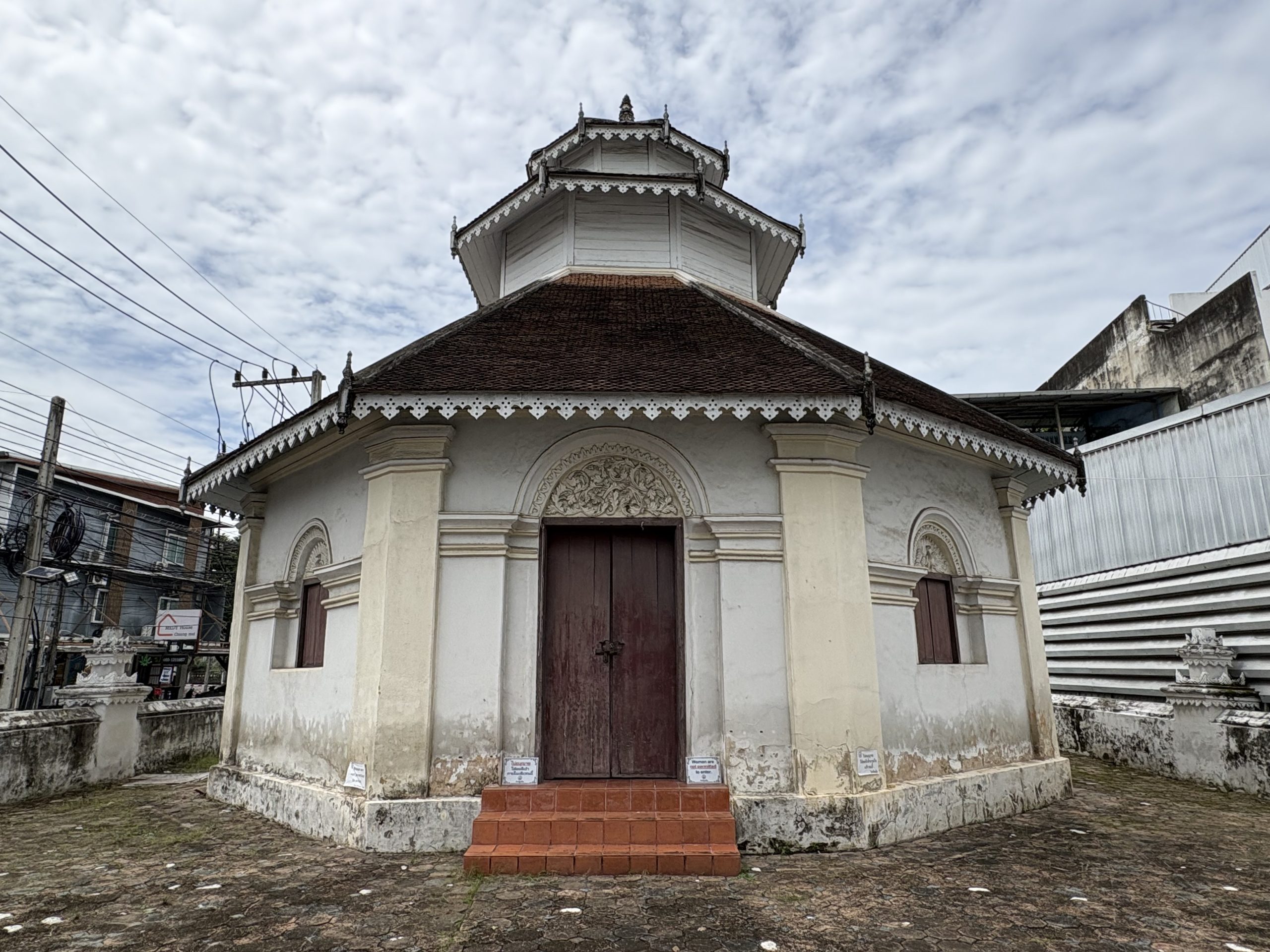 清迈 Wat Chiang Yuen 八角戒堂正面和斜向侧墙，棕红木门、白木叠顶檐饰、红台阶和街侧电线
