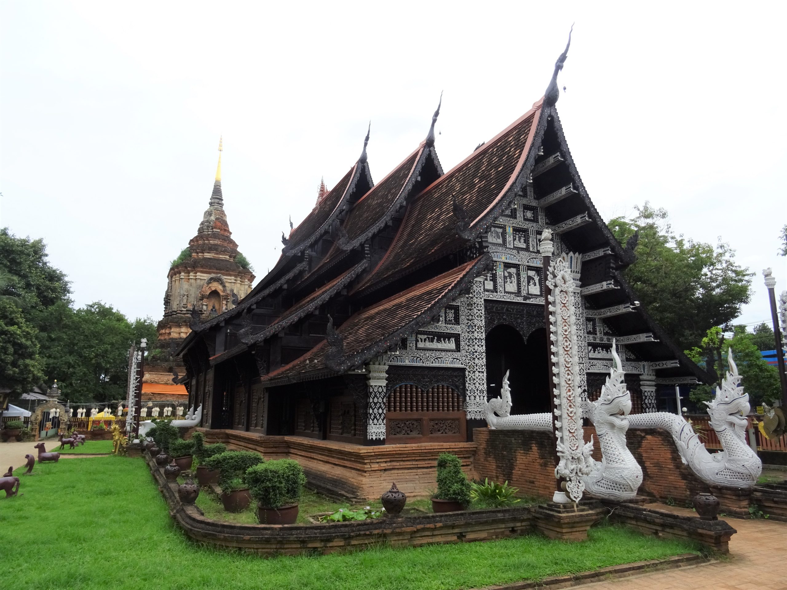 清迈 Wat Lok Moli 柚木殿的斜向正面，深色重叠山墙、红褐屋瓦、白色娜迦与侧后方砖塔