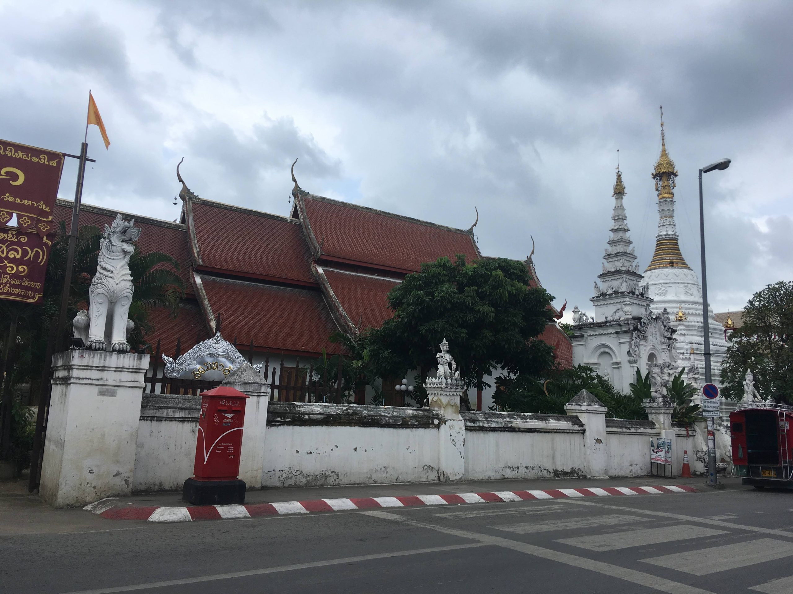 清迈 Wat Mahawan 白墙外的红邮筒与斑马线，院内露出红瓦殿顶和白色佛塔