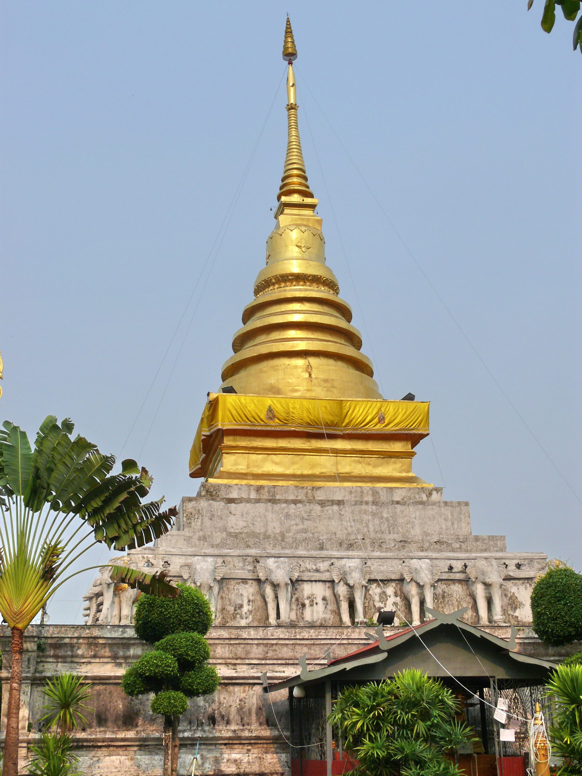 楠府象撑寺金色钟形塔,下方灰白象形装饰围绕方形塔座,前景有绿树与小棚。