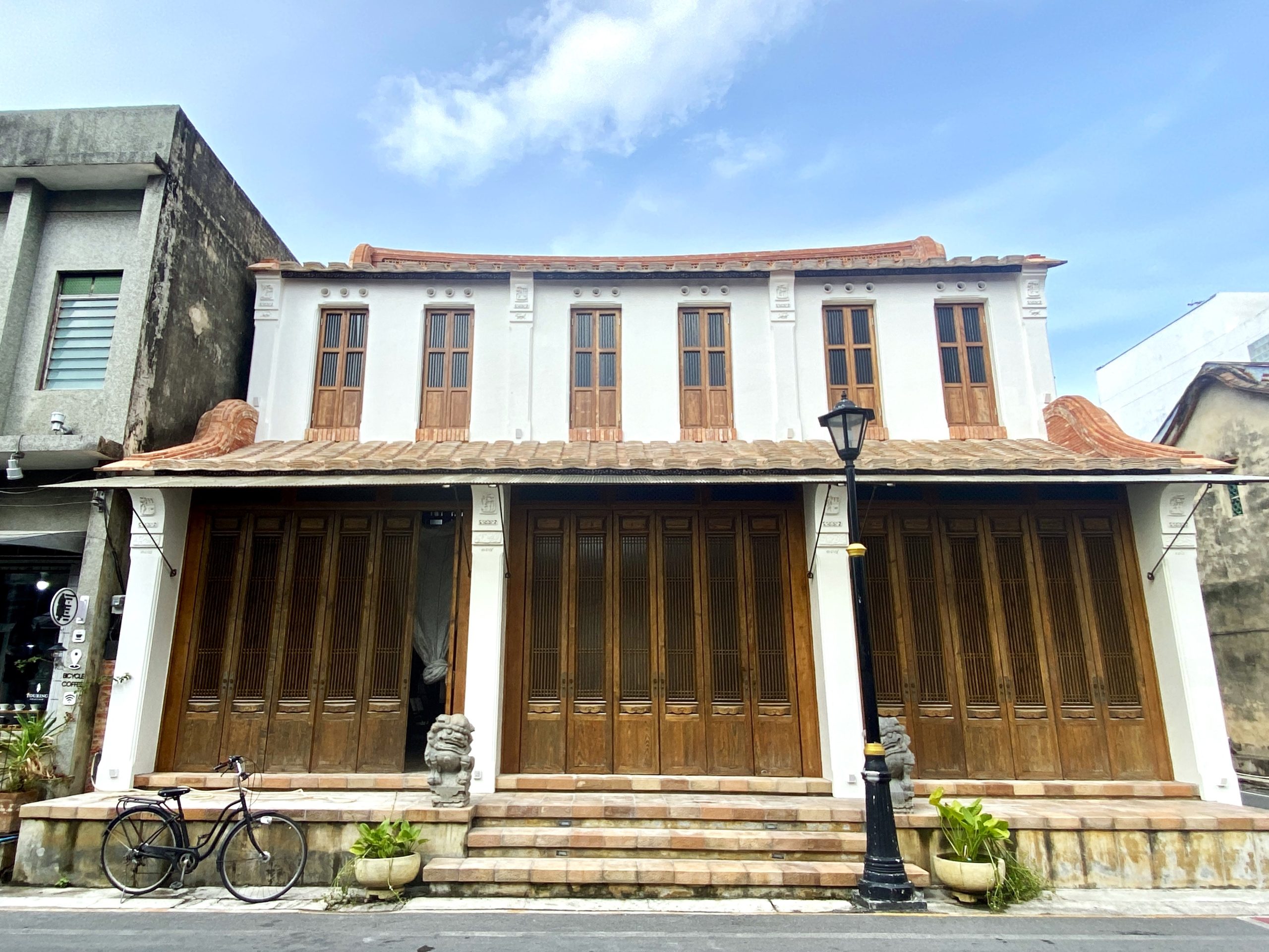 一座白色立面的临街建筑，连续木门和窗、红褐色装饰屋脊、台阶、黑色街灯和一辆自行车；两侧可见相邻建筑。