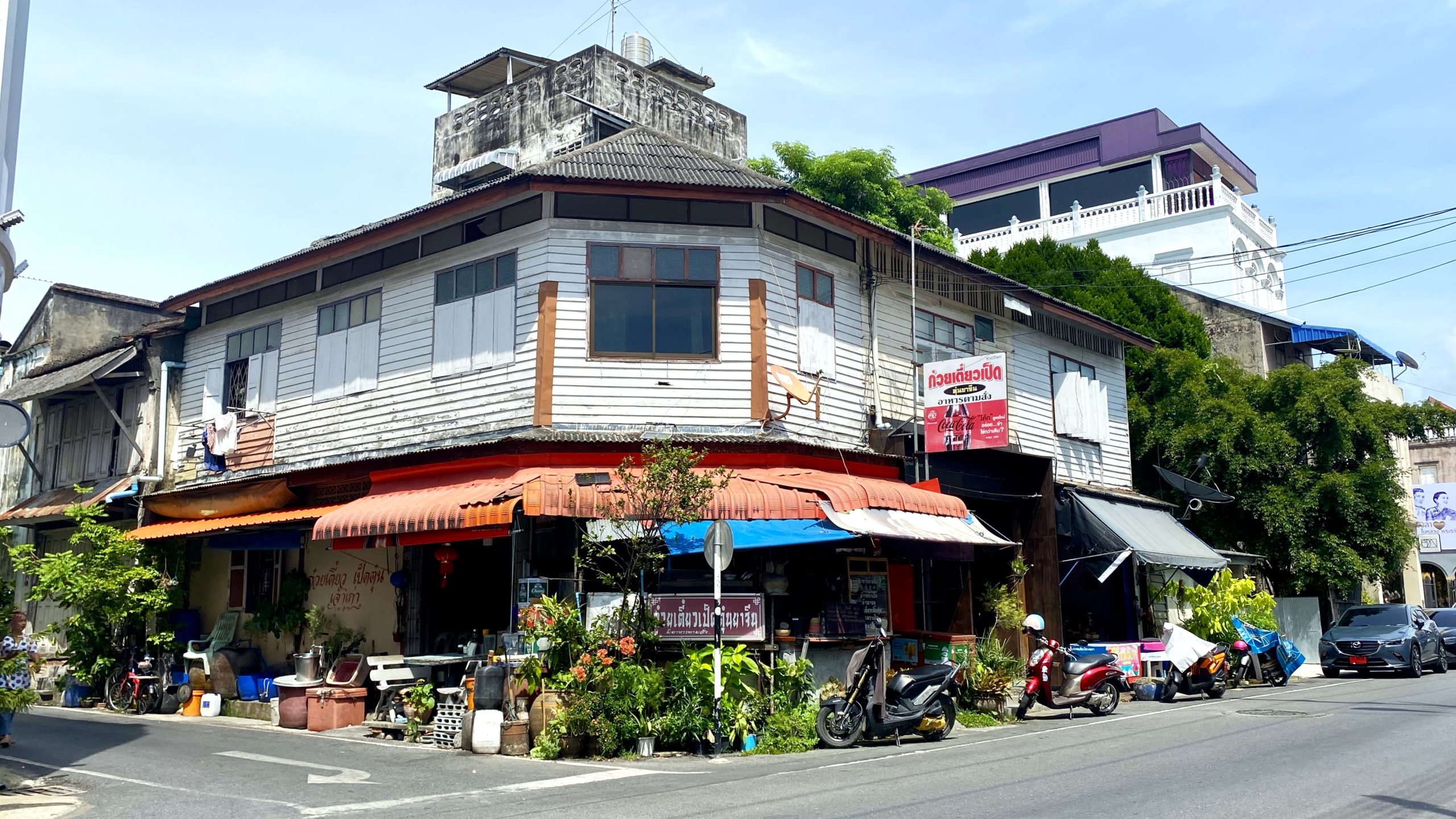 街边一排老式木板和混合材料建筑，底层有餐饮招牌、遮棚、盆栽、摩托车和道路交通；后方有较新的白色建筑。