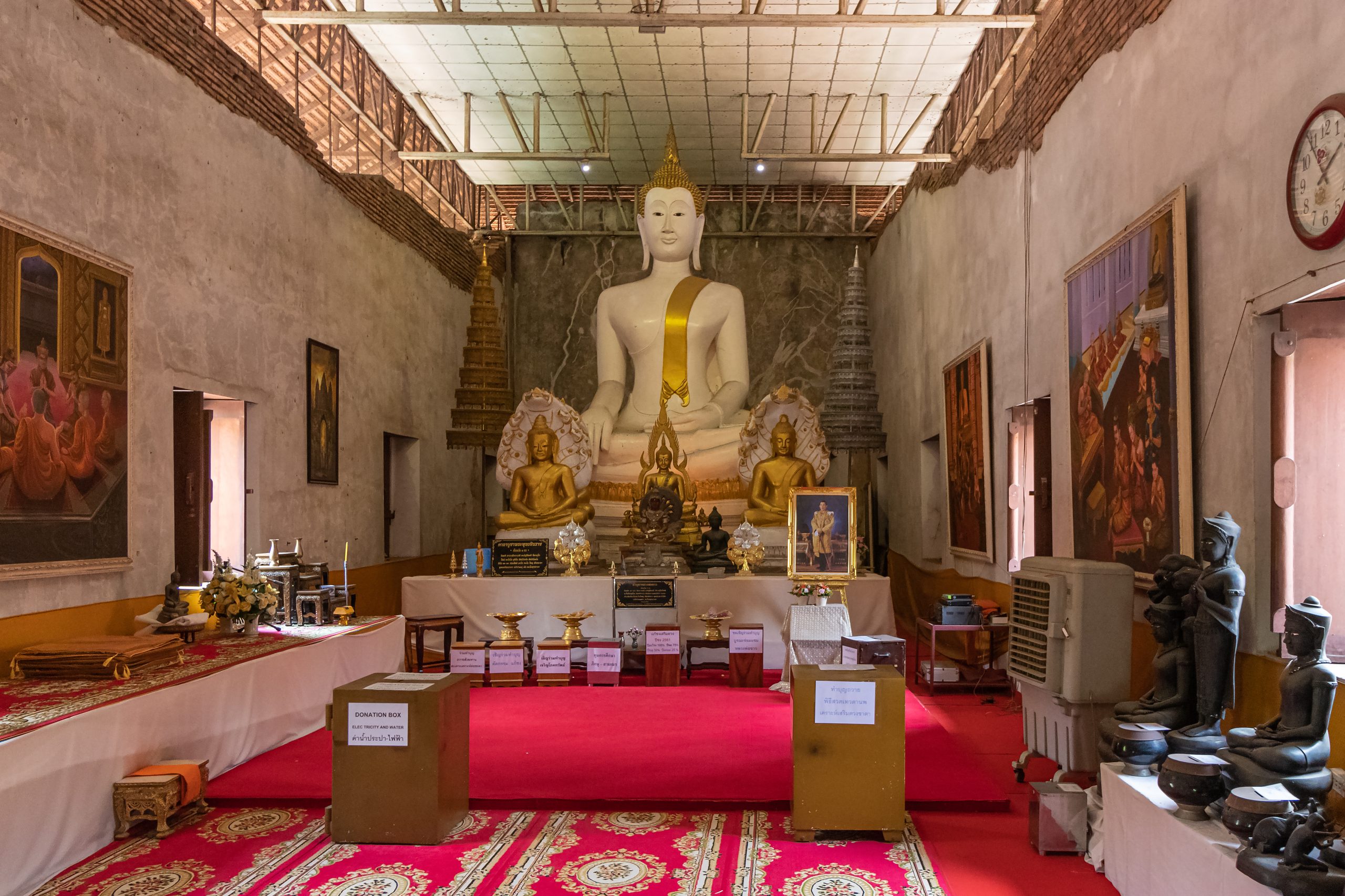 室内白色大坐佛披金色斜带,前有较小金佛,墙挂绘画,红地毯旁有供桌和募捐箱。